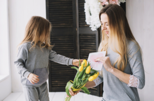 Aqui estão as opções de Alt Text para a imagem, adaptadas para o seu contexto de análise de mercado e Dia das Mães: Opção 1: Foco em Contexto de Consumo e E-commerce "Criança entregando um buquê de tulipas amarelas e um cartão feito à mão com desenho de coração para sua mãe, ilustrando o comportamento de compra e as categorias de destaque no Dia das Mães.