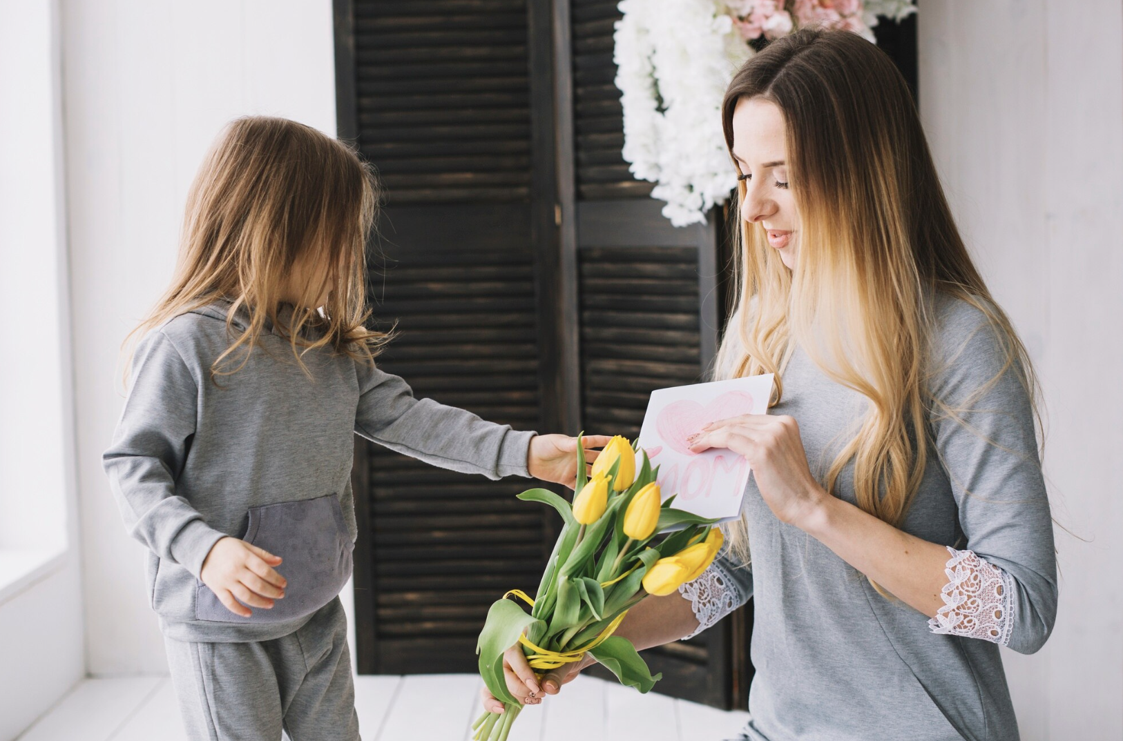 Aqui estão as opções de Alt Text para a imagem, adaptadas para o seu contexto de análise de mercado e Dia das Mães: Opção 1: Foco em Contexto de Consumo e E-commerce "Criança entregando um buquê de tulipas amarelas e um cartão feito à mão com desenho de coração para sua mãe, ilustrando o comportamento de compra e as categorias de destaque no Dia das Mães.