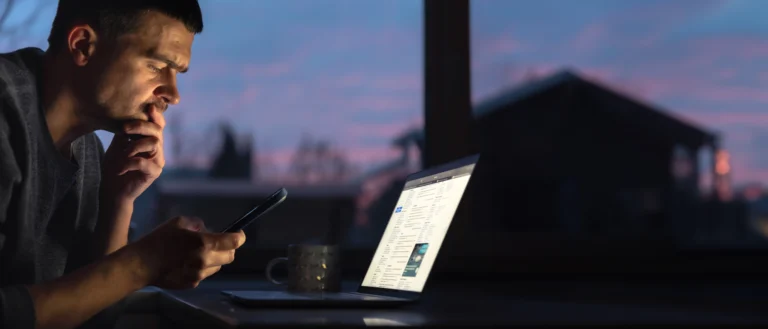 Close-up de um homem de perfil com expressão pensativa, deitado à noite, olhando para um smartphone na mão direita e com a mão esquerda no queixo. À frente, um laptop
