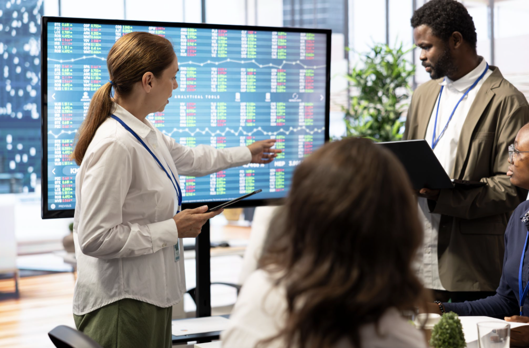 Uma executiva de perfil aponta para uma grande tela digital exibindo gráficos financeiros, planilhas analíticas de mercado e tabelas com dados de desempenho e métricas de vendas, durante uma reunião de alinhamento com sua equipe.