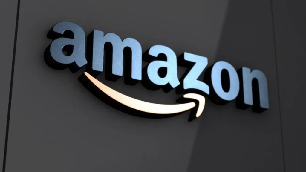 O logotipo da Amazon, em material metálico azul prateado com brilho e contornos azuis mais escuros, está afixado em uma parede cinza escura texturizada. A famosa seta "sorriso" da Amazon está logo abaixo, iluminada por uma luz branca quente que projeta um brilho suave e dourado para baixo na parede.