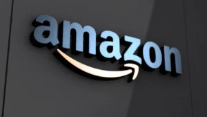 O logotipo da Amazon, em material metálico azul prateado com brilho e contornos azuis mais escuros, está afixado em uma parede cinza escura texturizada. A famosa seta "sorriso" da Amazon está logo abaixo, iluminada por uma luz branca quente que projeta um brilho suave e dourado para baixo na parede.