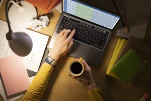 Visão aérea de uma pessoa digitando em um laptop em uma mesa de madeira iluminada por uma luminária, segurando uma caneca amarela com café preto ao lado de livros e papéis com anotações.